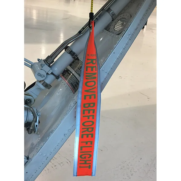Red Remove Before Flight tag hanging from an aircraft control surface assembly, warning to remove before flight to ensure safety